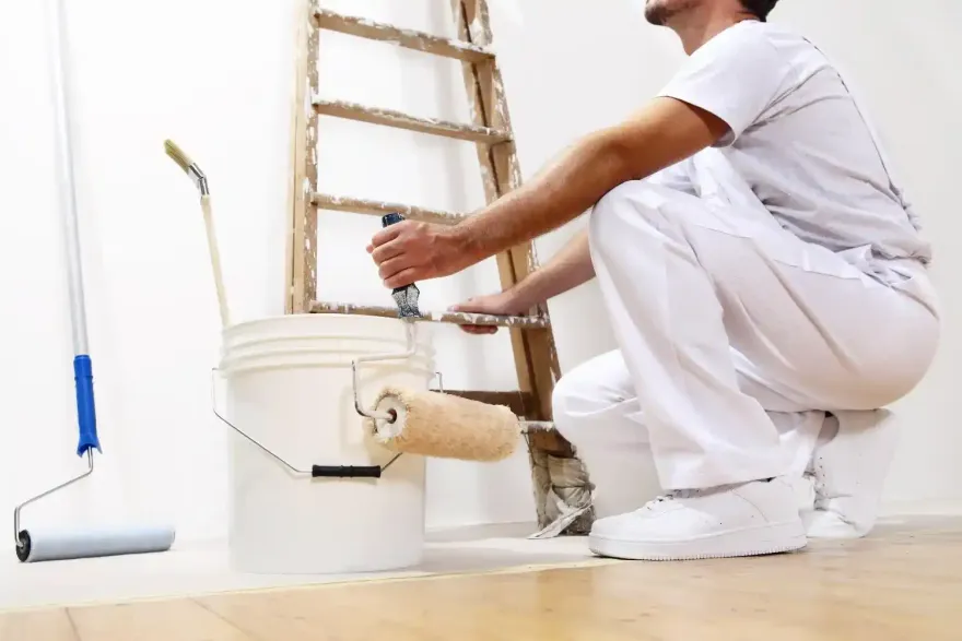 Una persona dipinge un secchio bianco su un pavimento di legno accanto a una scala e un rullo per dipingere.
