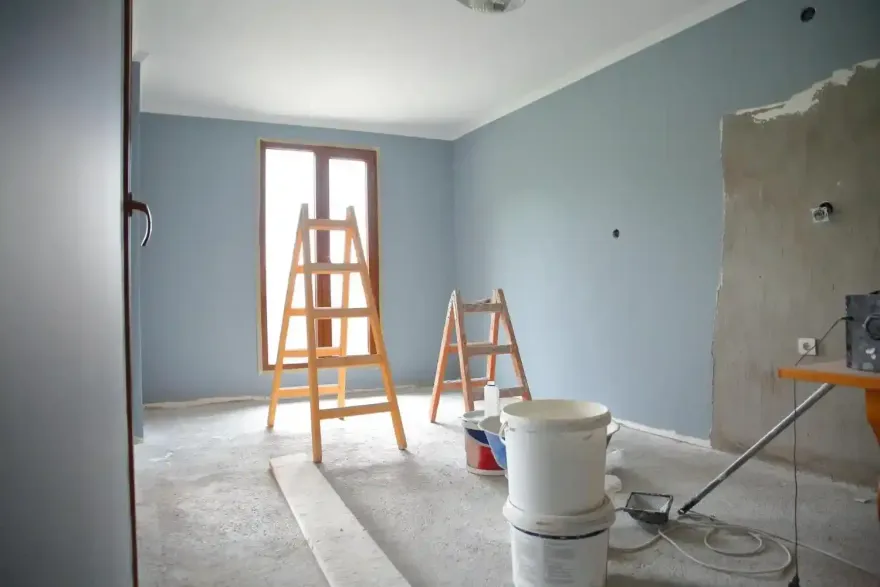 Stanza parzialmente ristrutturata con pareti blu, materiale per dipingere e due scalette su un pavimento di cemento.