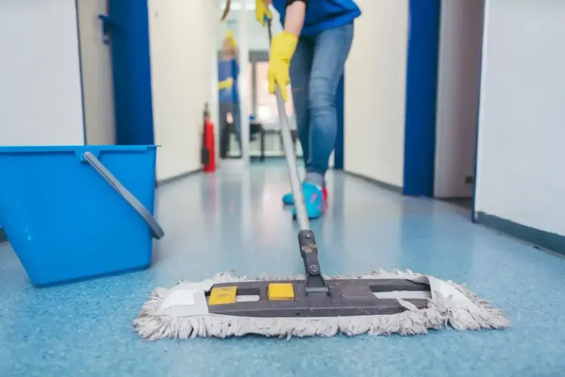Una persona sta lavando un pavimento blu in un corridoio, con un secchio giallo nelle vicinanze.