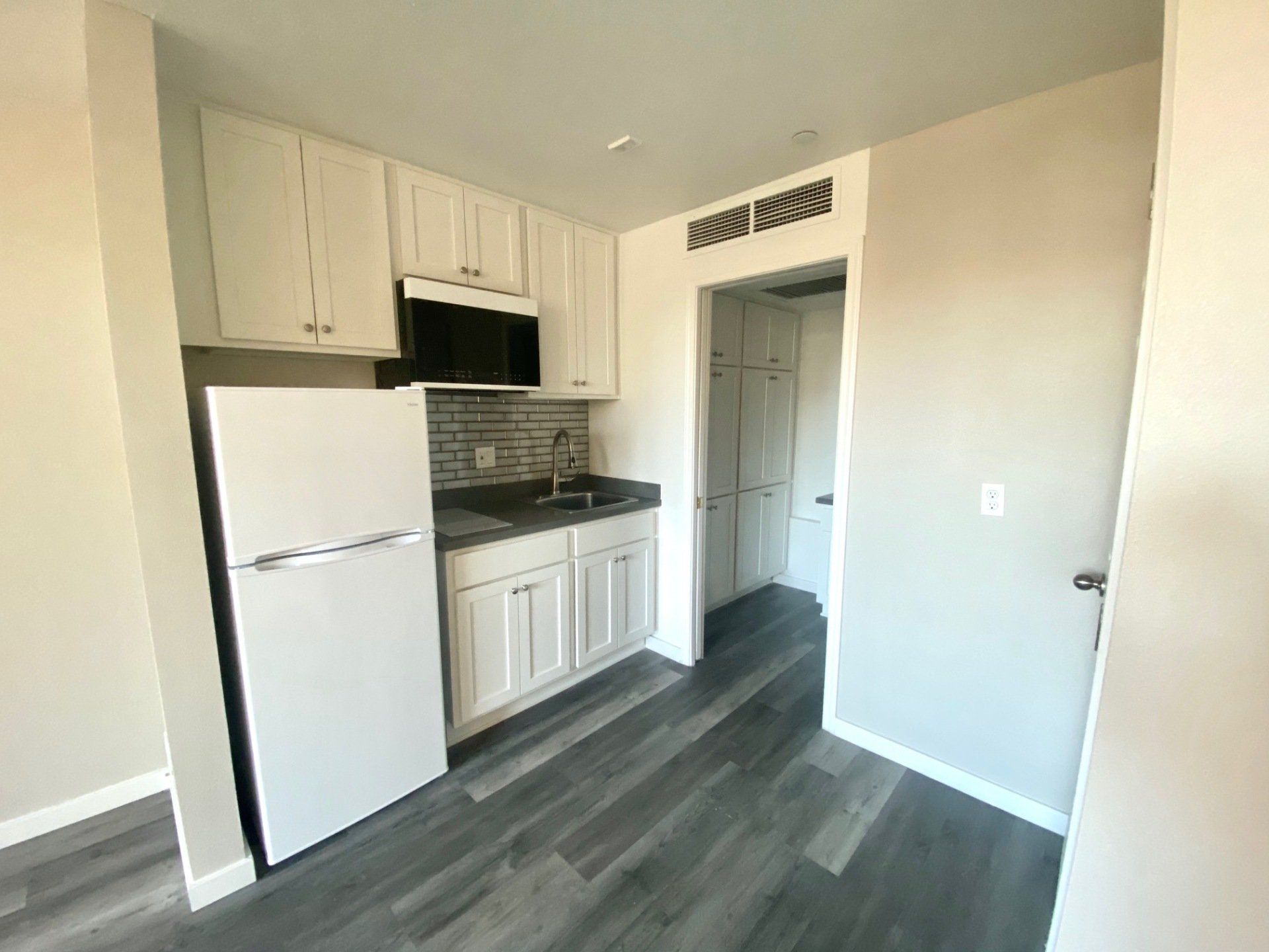 Updated studio kitchen with grey flooring, white cabinets, grey countertops, black microwave, white two electric burner stove, and white refrigerator.