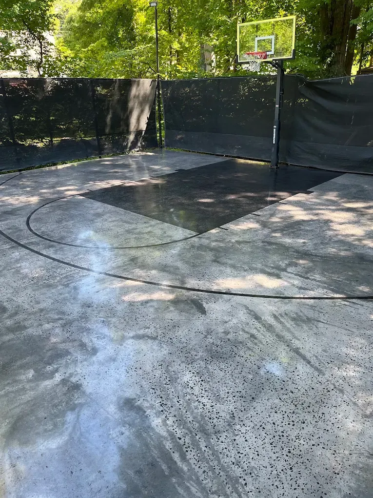 Outdoor basketball half-court with a hoop, black fence, and wet concrete surface surrounded by trees.
