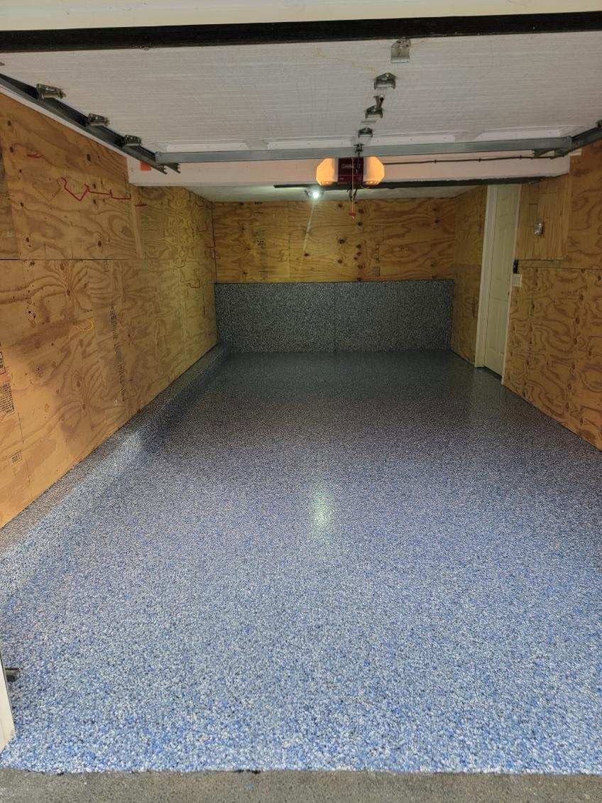Empty garage with a speckled blue floor and plywood-lined walls, viewed from the open entrance