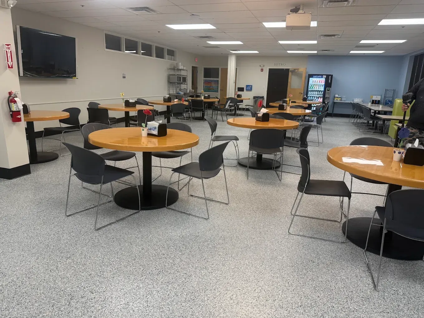 Empty cafeteria with round wooden tables, black chairs, vending machines, and a TV on the wall.