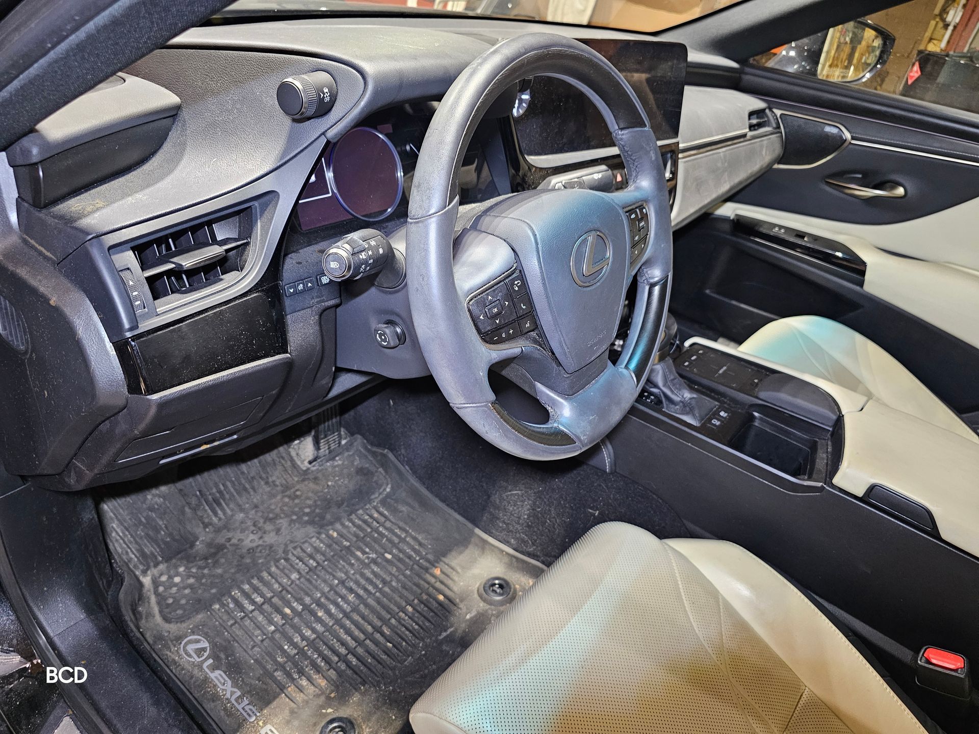 Interior view of a Lexus dashboard, steering wheel, and seats. Black and cream color scheme, dirty floor mats.