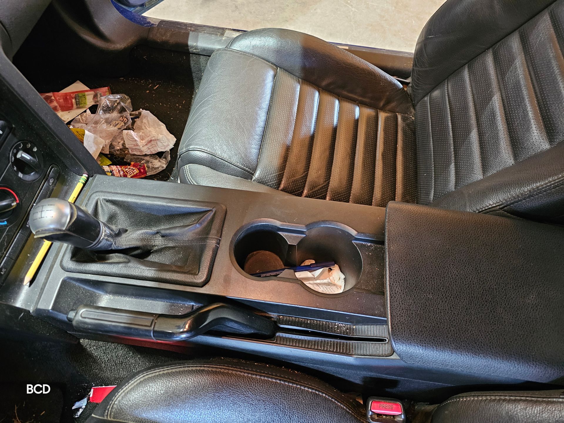 Interior car view of black gearshift, cup holders, and leather seats. Debris in front seat.