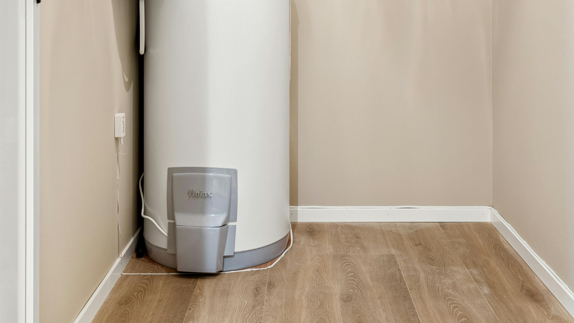 A white electric water heater stands in the corner of a room with light beige walls and light brown wood-look flooring.