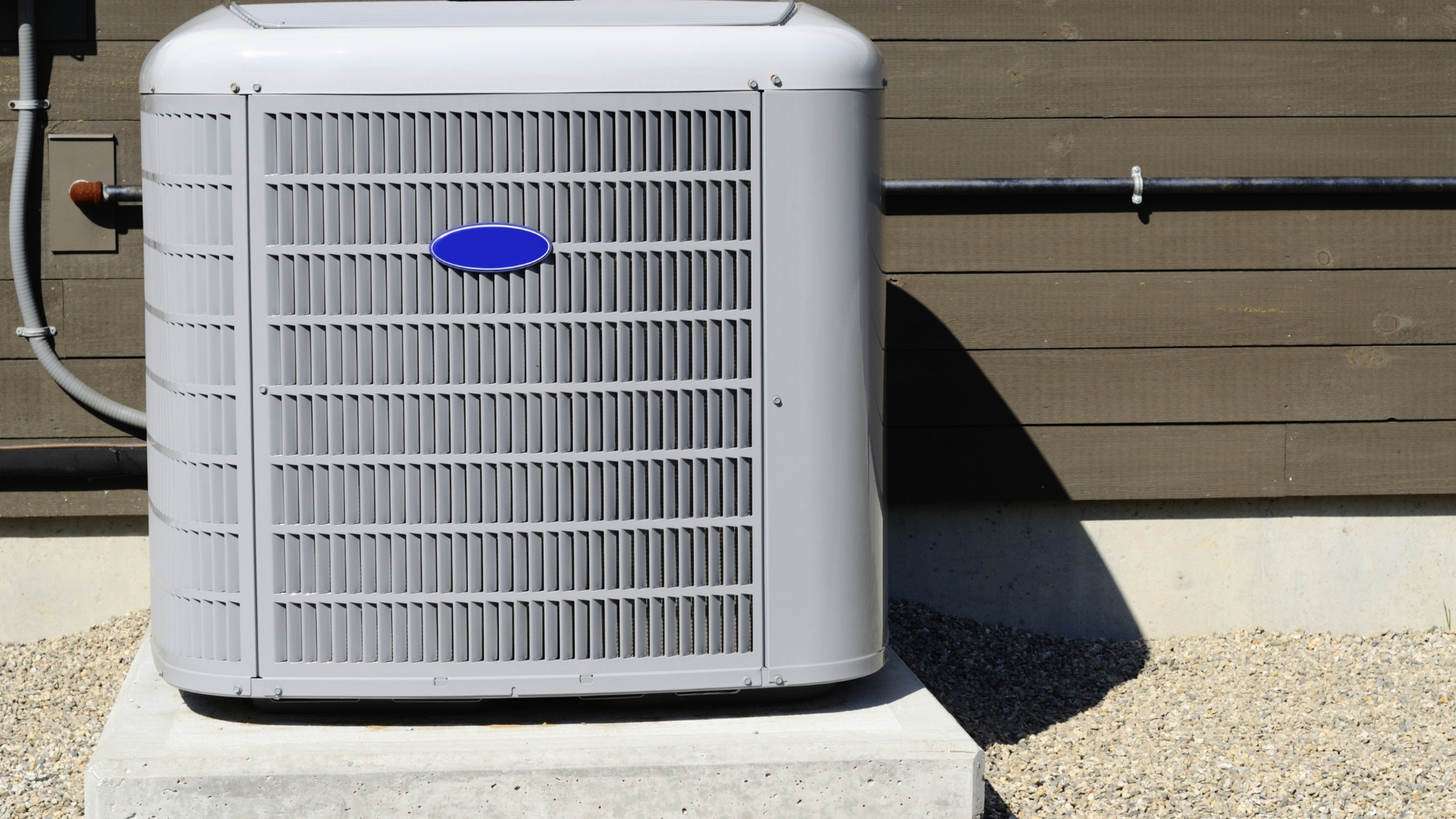A white outdoor HVAC condensing unit sitting on a concrete pad against a brown wooden wall.