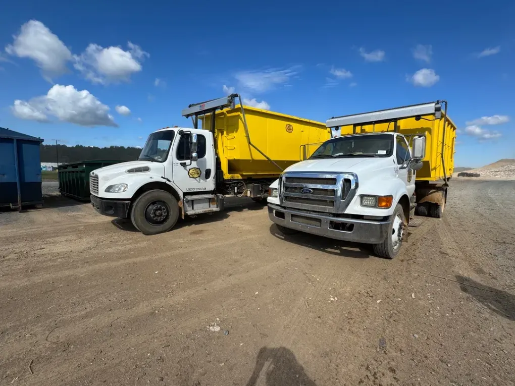 Two white dump trucks with yellow beds parked on a gravel lot under a blue sky.