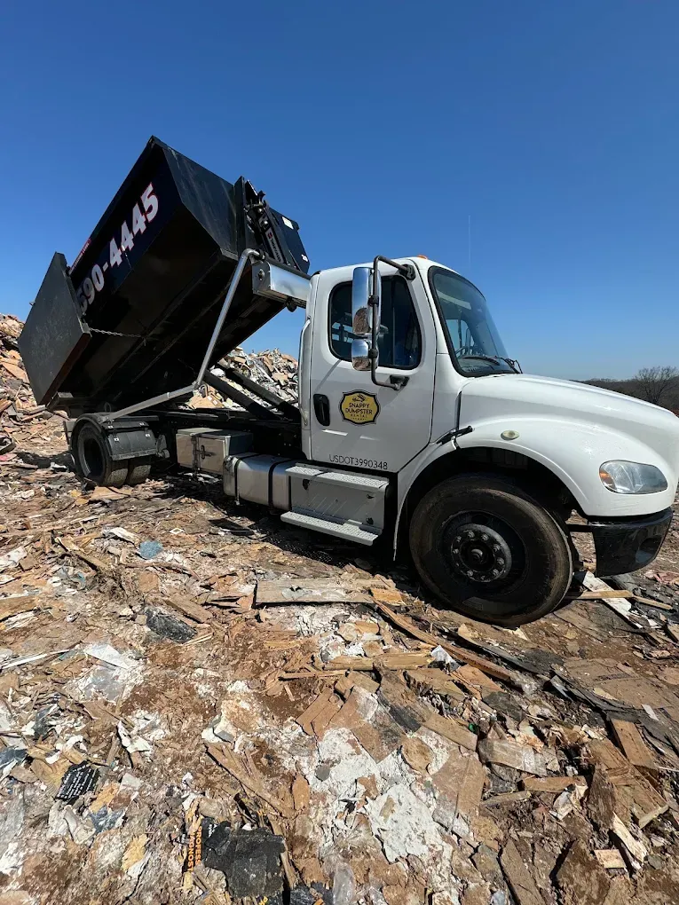 White roll-off truck on a rocky slope under a clear blue sky, truck bed raised and tilted.