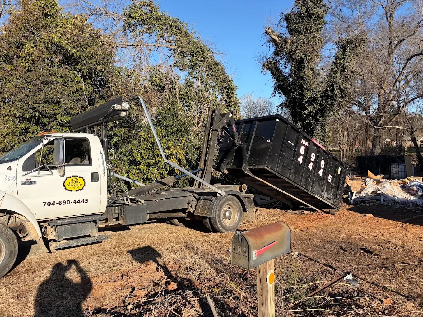 Hooklift truck lifting a black dumpster in a wooded area with debris and a mailbox nearby