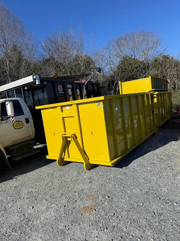 Yellow construction dumpster beside a roll-off truck on a gravel lot under a clear blue sky