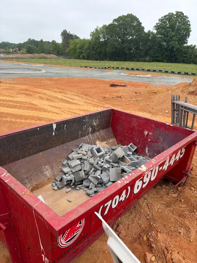 Red construction dumpster filled with gray concrete on an orange dirt site near trees and open ground