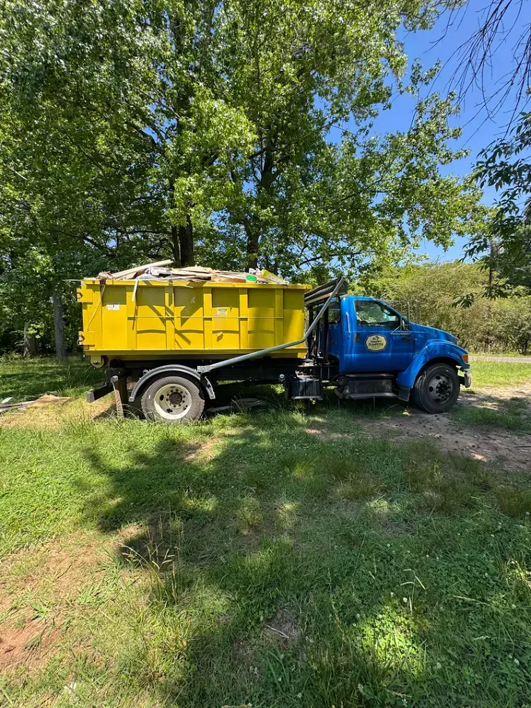 Blue roll-off truck with a yellow bed parked on grass under leafy trees in sunlight