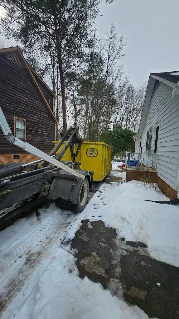 Snowy driveway beside houses with a yellow dumpster and a small excavator at left