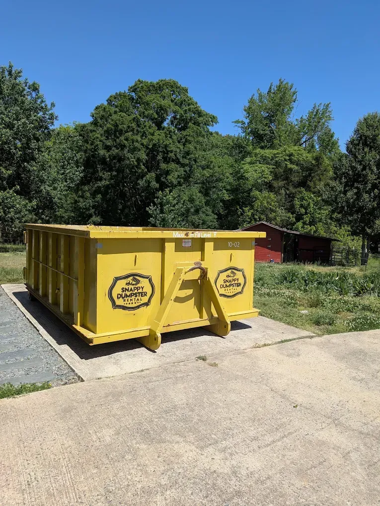 Yellow dumpster outdoors beside a gravel path, with trees and a small red shed in the background
