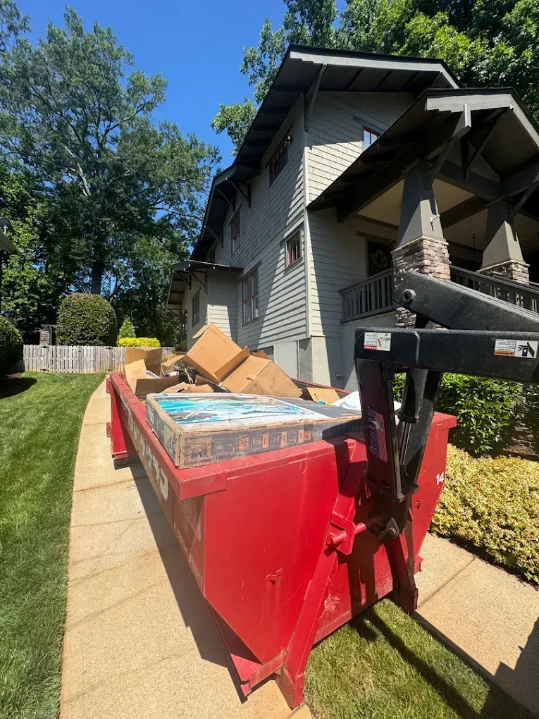 Red dumpster full of debris beside a gray house in a sunny yard