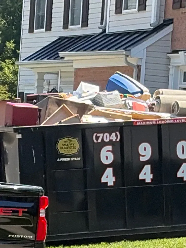 Construction dumpster overflowing with debris in front of a house, with a pickup truck beside it
