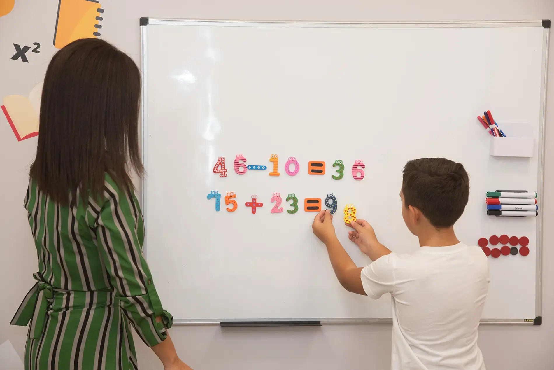 Μια γυναίκα στέκεται δίπλα σε ένα αγόρι που γράφει σε έναν πίνακα.