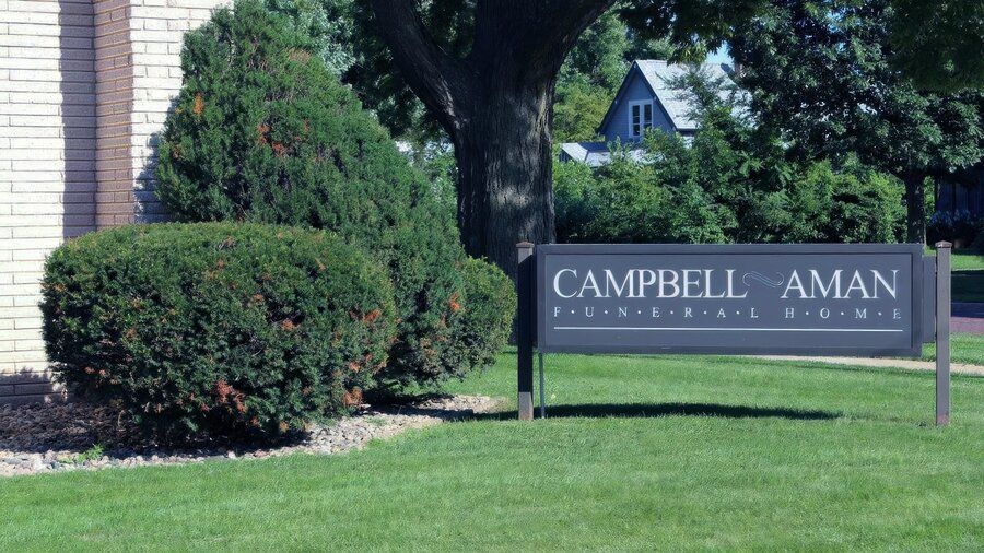 Campbell-Aman Funeral Home dark grey outdoor sign set on a green lawn with manicured bushes and trees.