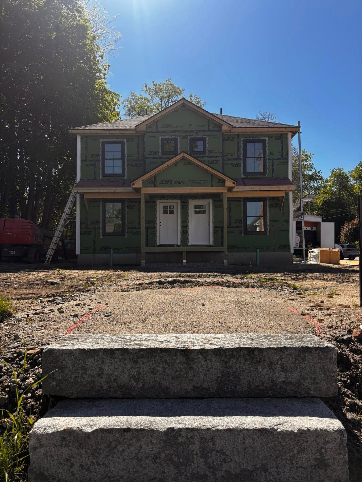 Green two-story house under construction with gravel front yard and concrete steps in sunlight
