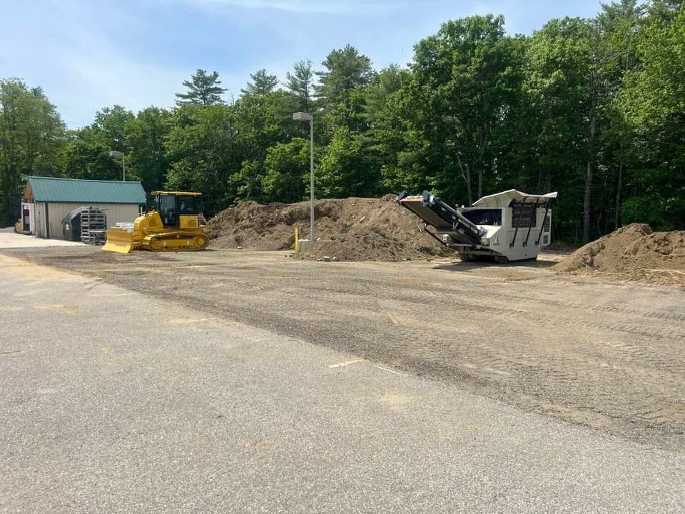 Construction yard with a bulldozer, dump truck, and dirt piles beside a wooded area