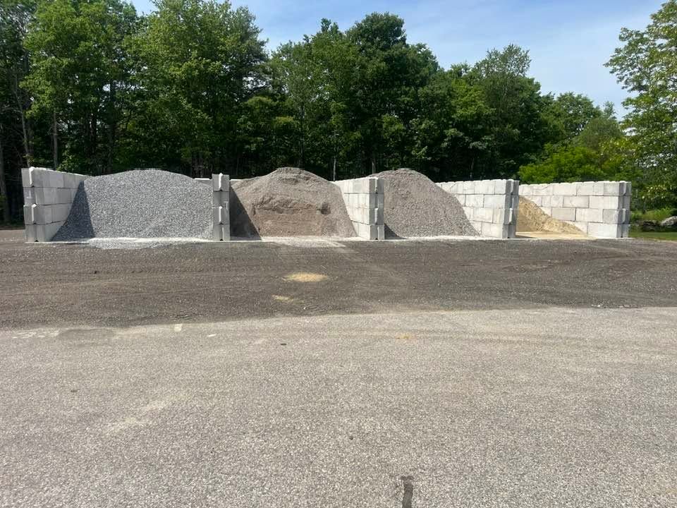 Gravel and sand piles beside a white block wall in a wooded outdoor storage yard