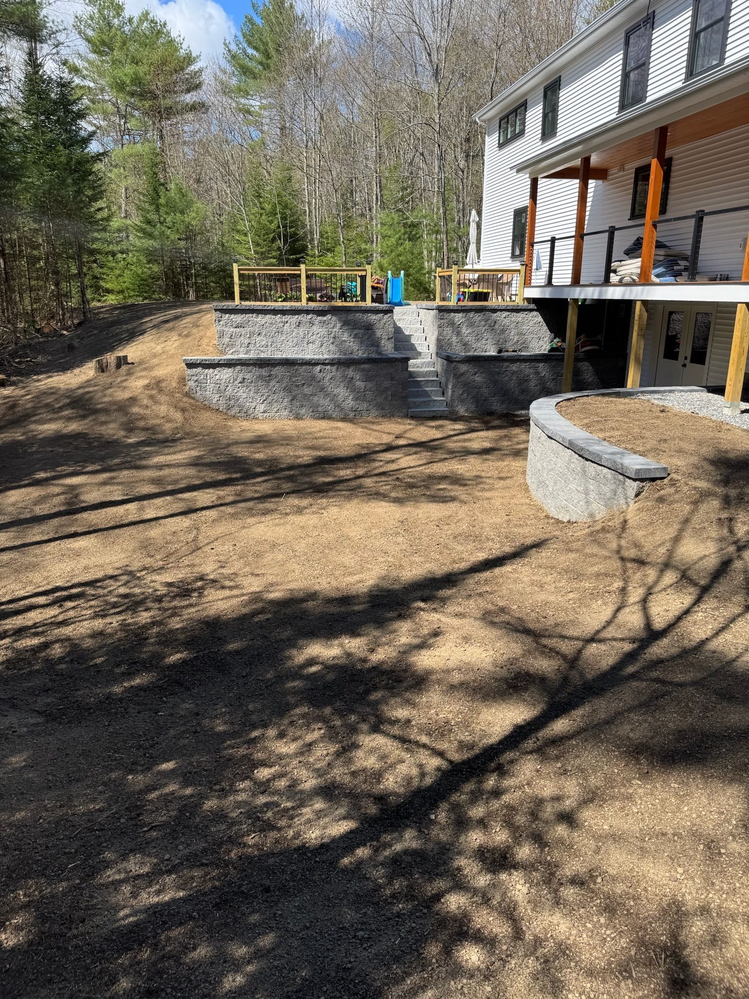 Outdoor patio with stone retaining walls, deck, and trees in a sunny backyard