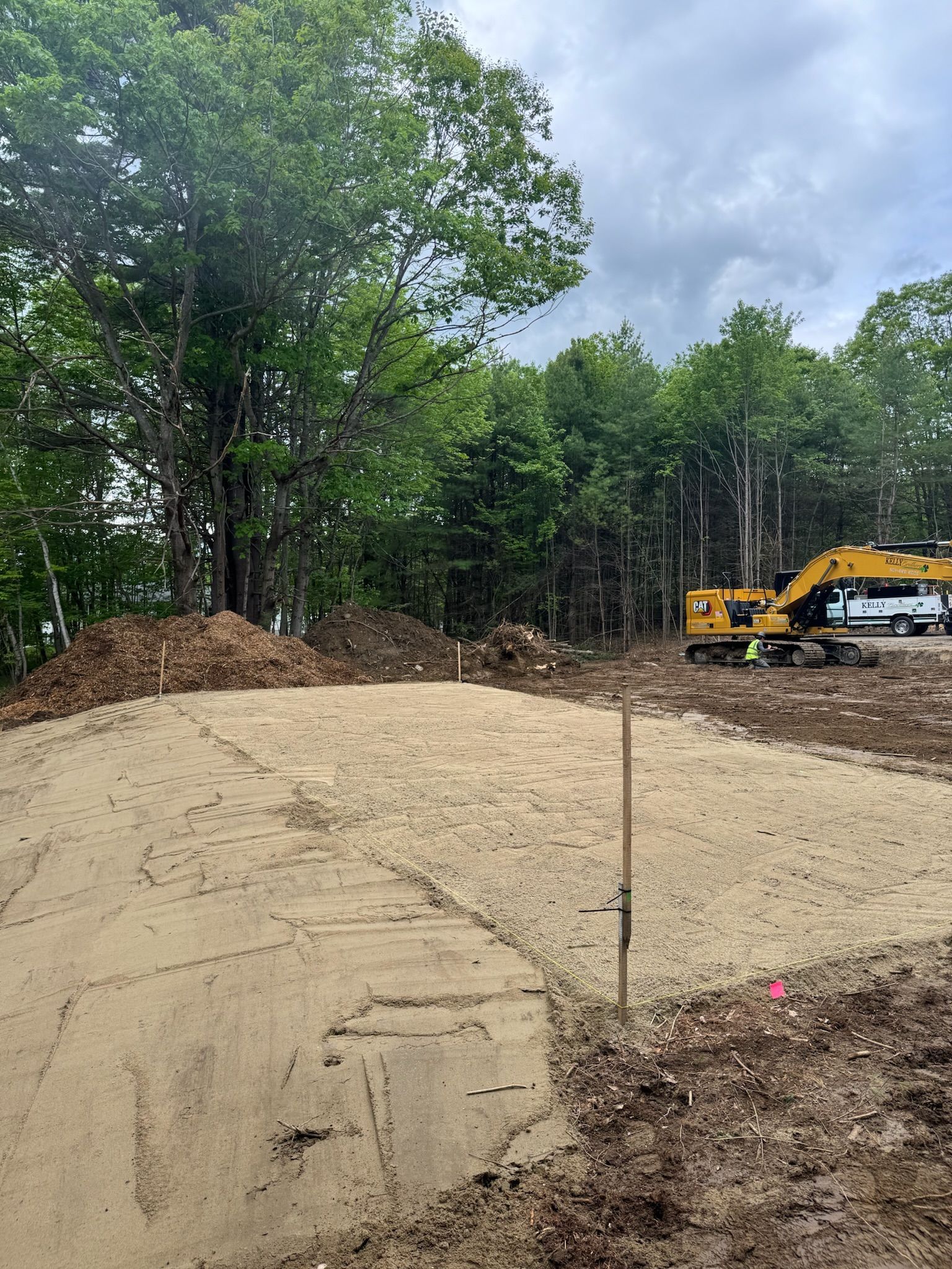 Cleared sandy lot with dirt piles, trees in background, and a yellow excavator at right under cloudy sky