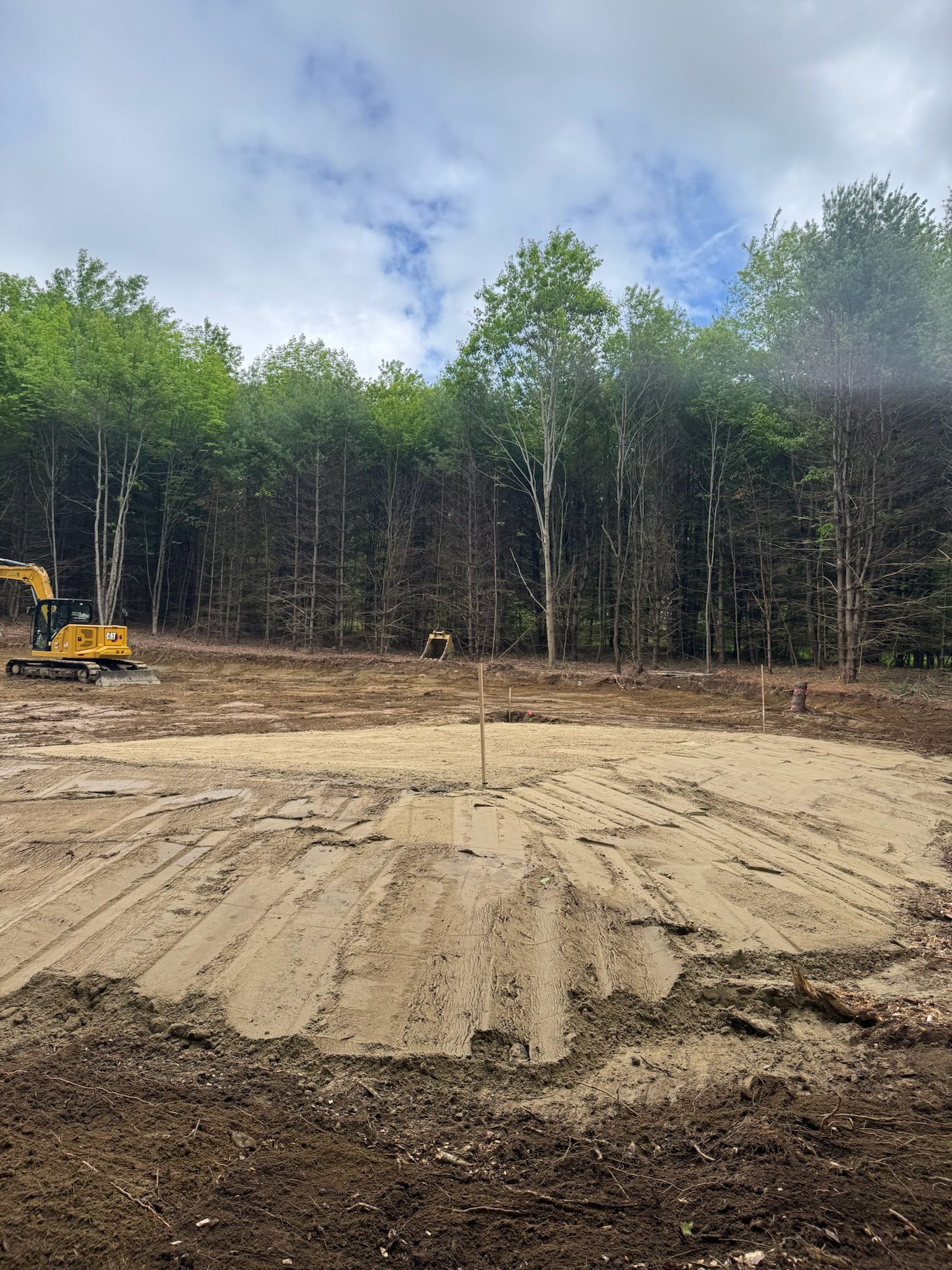 Cleared dirt lot with a yellow excavator and trees under a cloudy sky