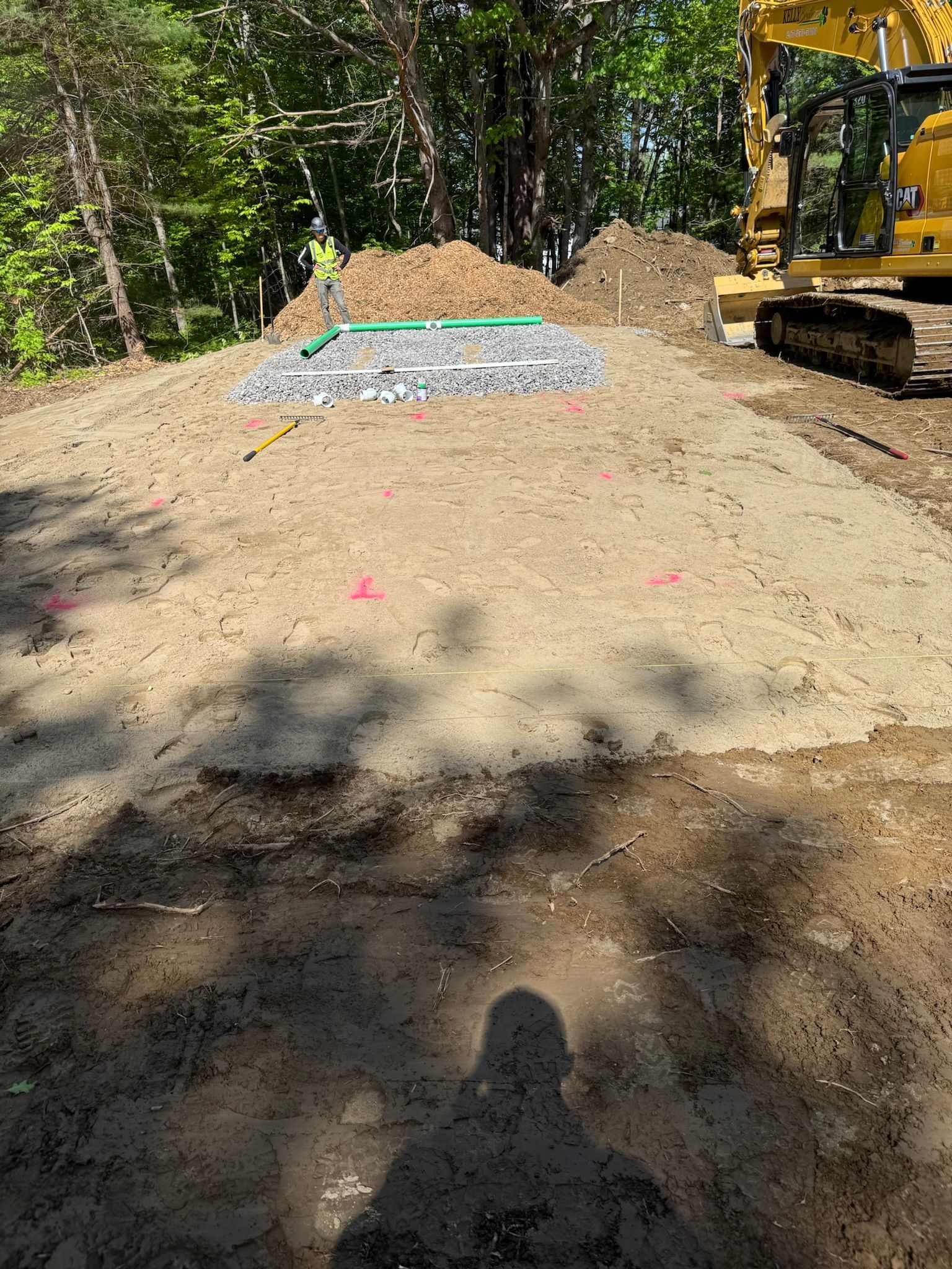 Large dirt pile at a wooded construction site beside an excavator arm and gravel mound