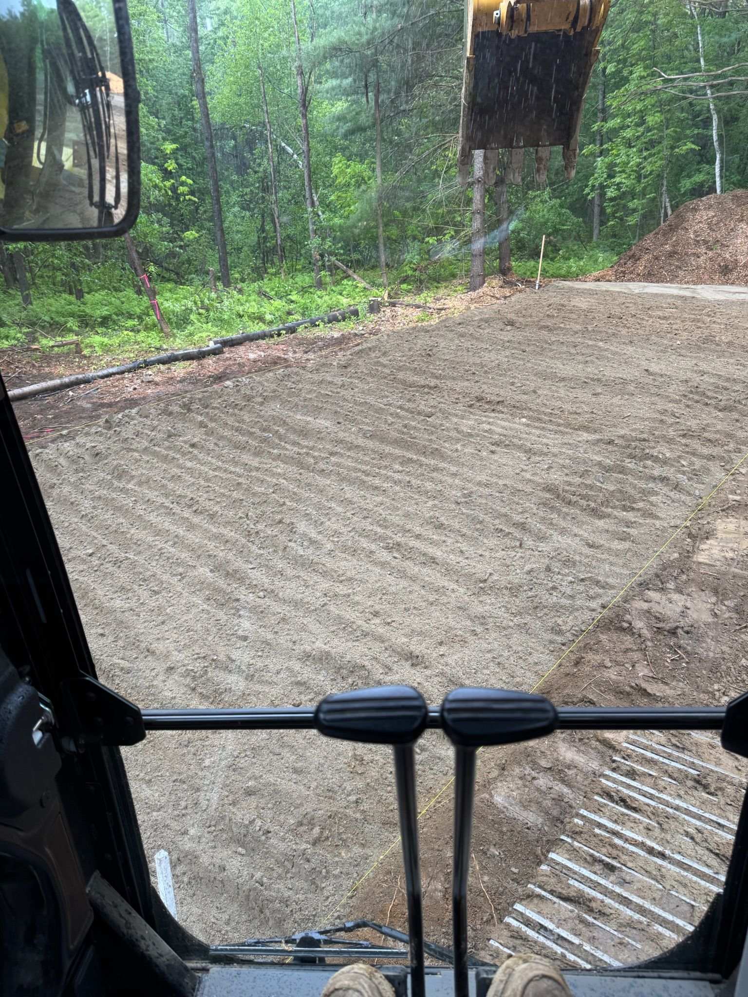 View from a construction vehicle over a freshly graded gravel road in a wooded area, with excavator arms visible.