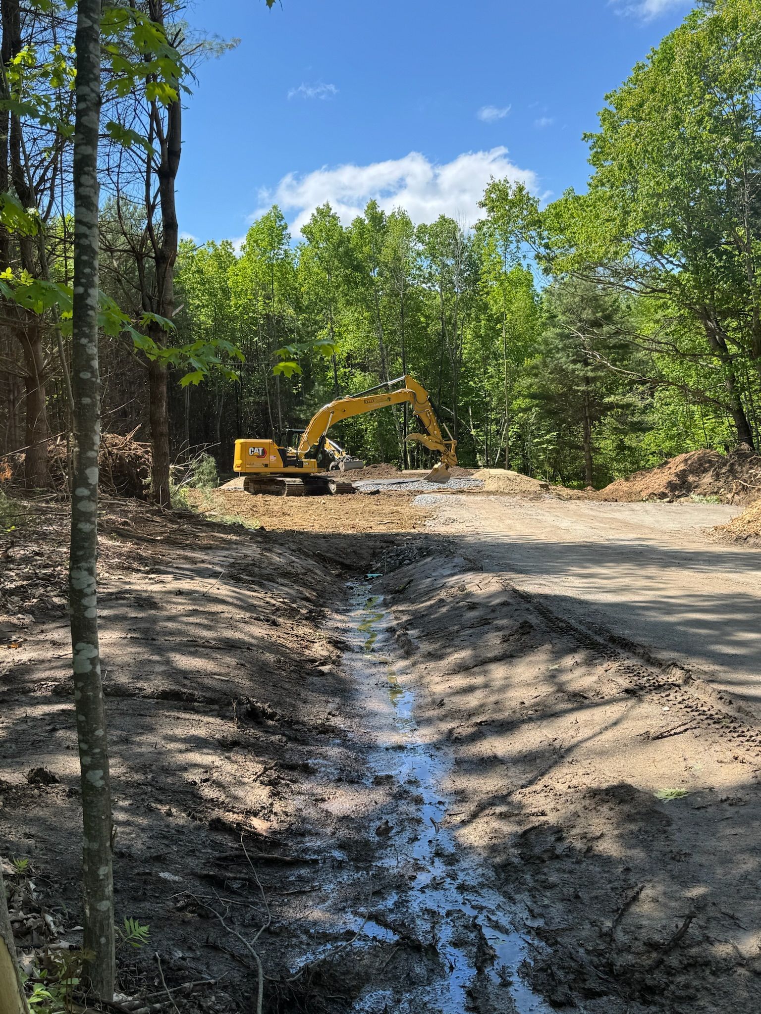 Yellow excavator on muddy dirt road in a wooded area with a small stream in the foreground