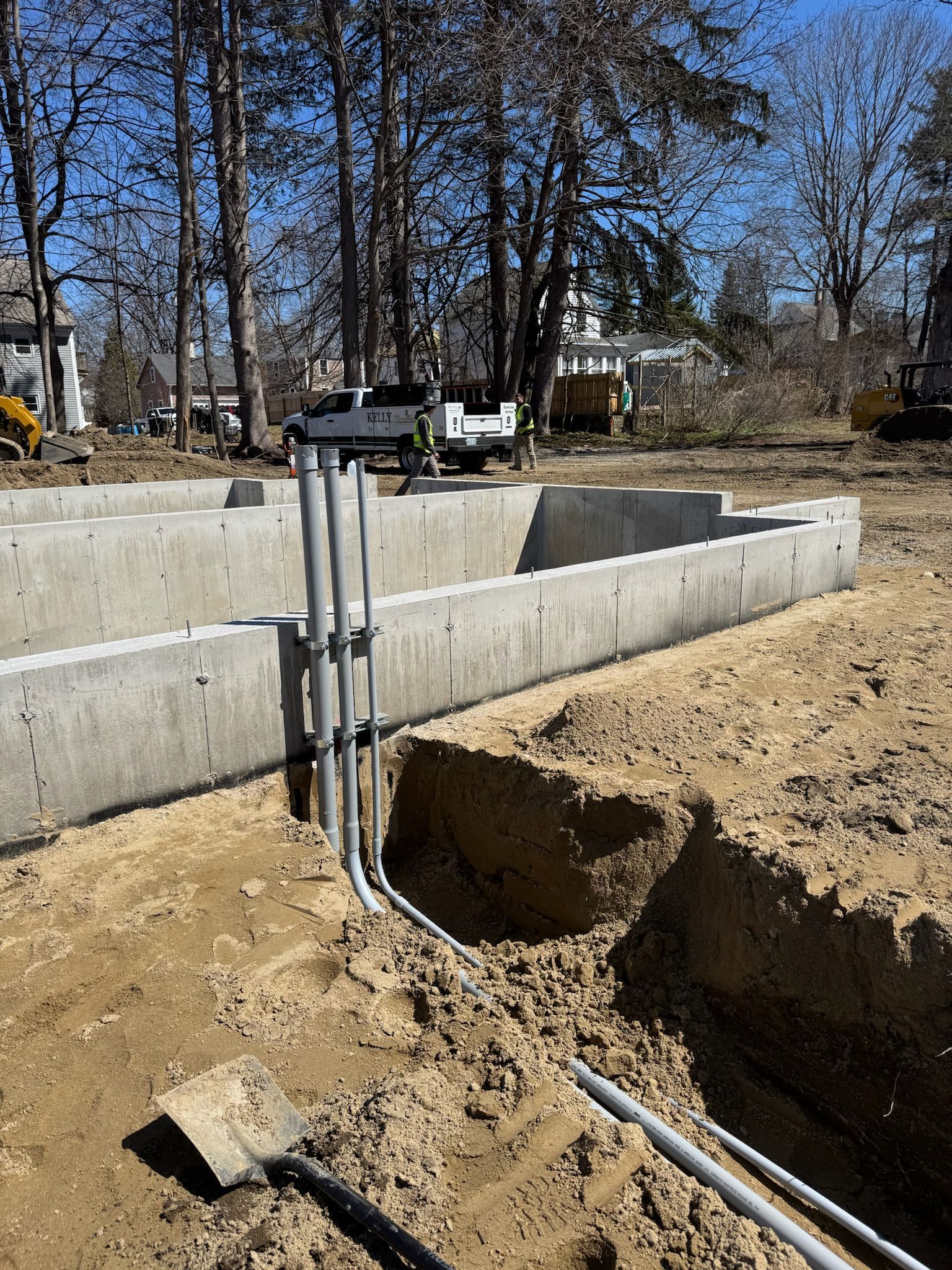 Concrete foundation trench at a construction site with exposed pipes and dirt under bare trees.