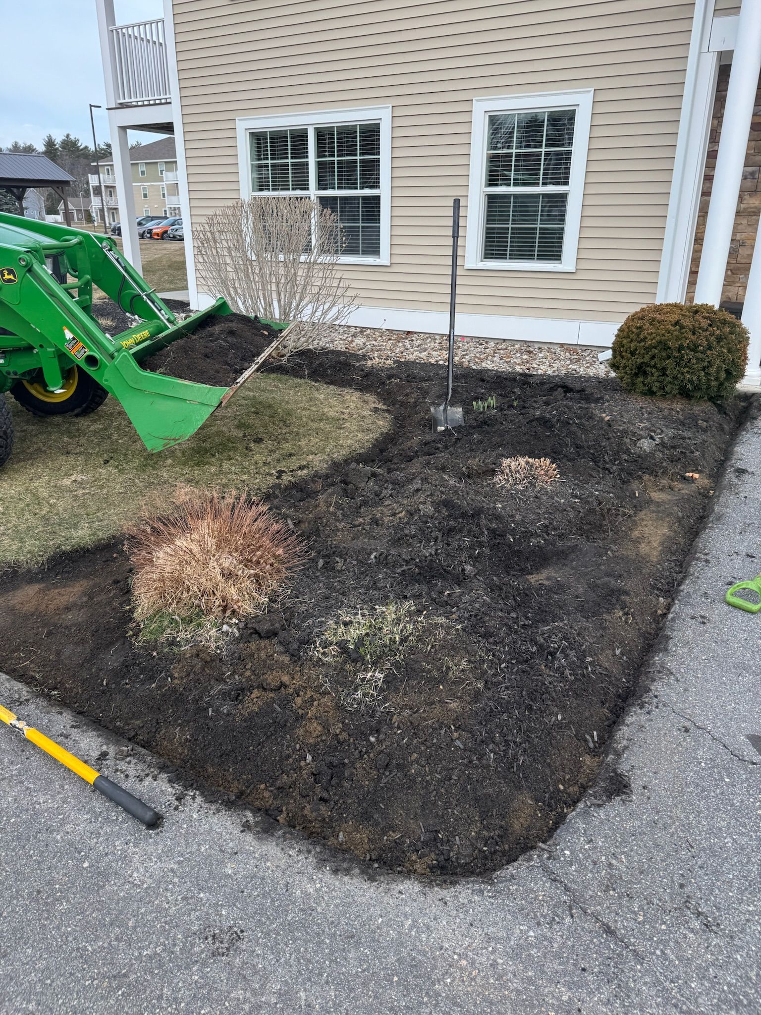 Landscaped bed being cleaned with a green tractor bucket beside a tan house and windows