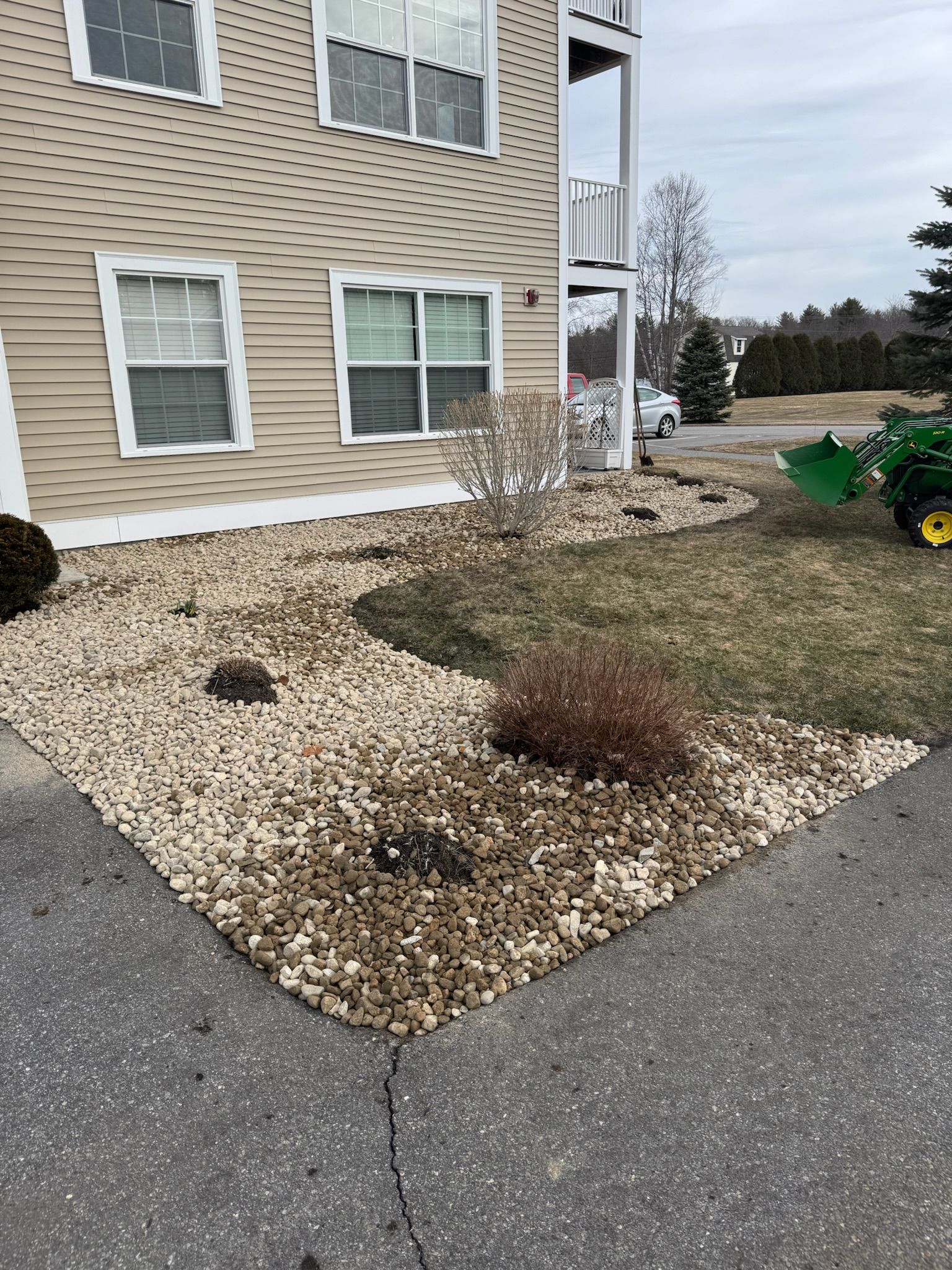 Apartment building corner with rock mulch landscaping, small shrubs, and a patchy winter lawn beside a sidewalk.