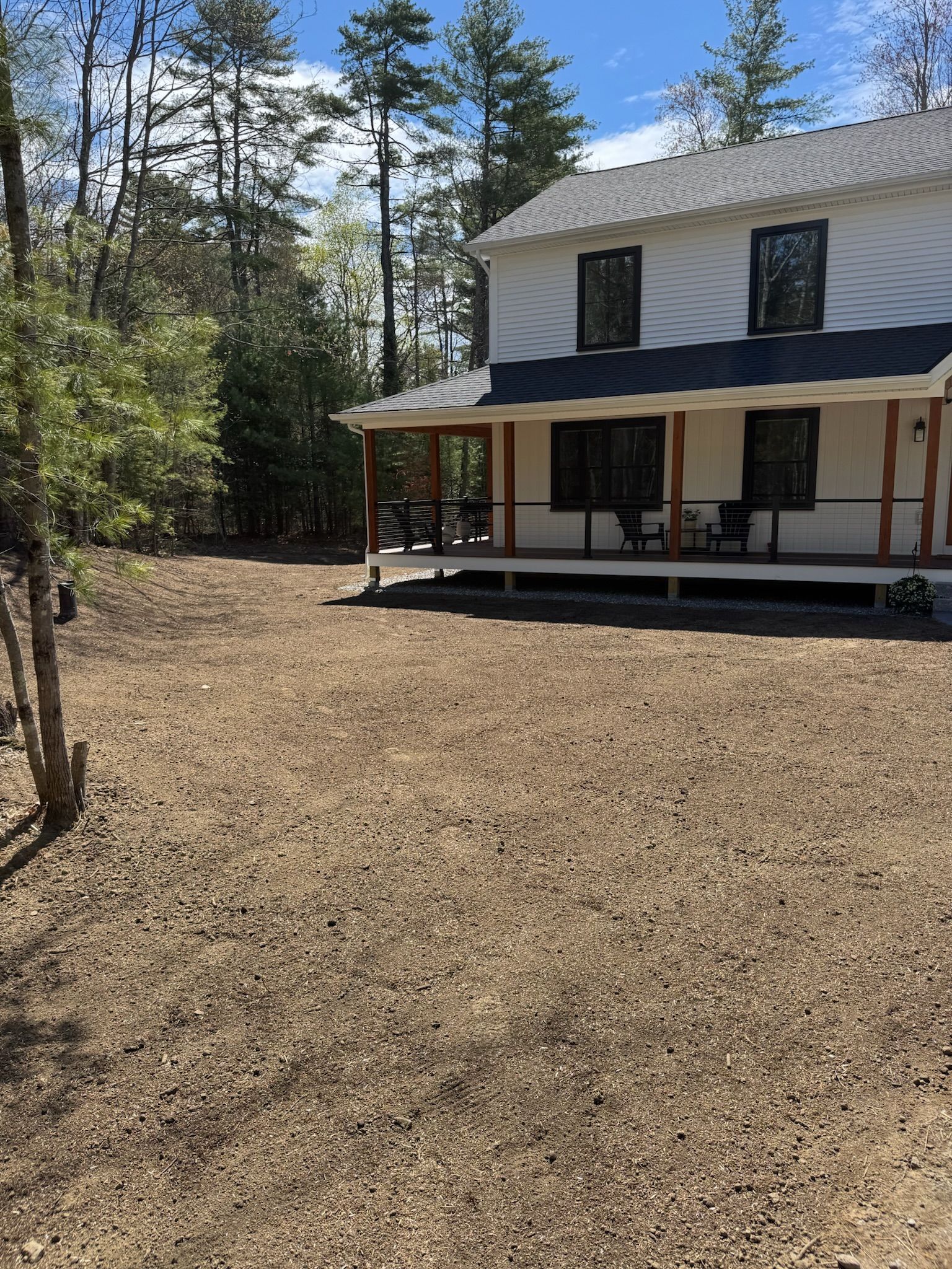 Two-story house with a covered porch beside a gravel yard and trees under a blue sky.