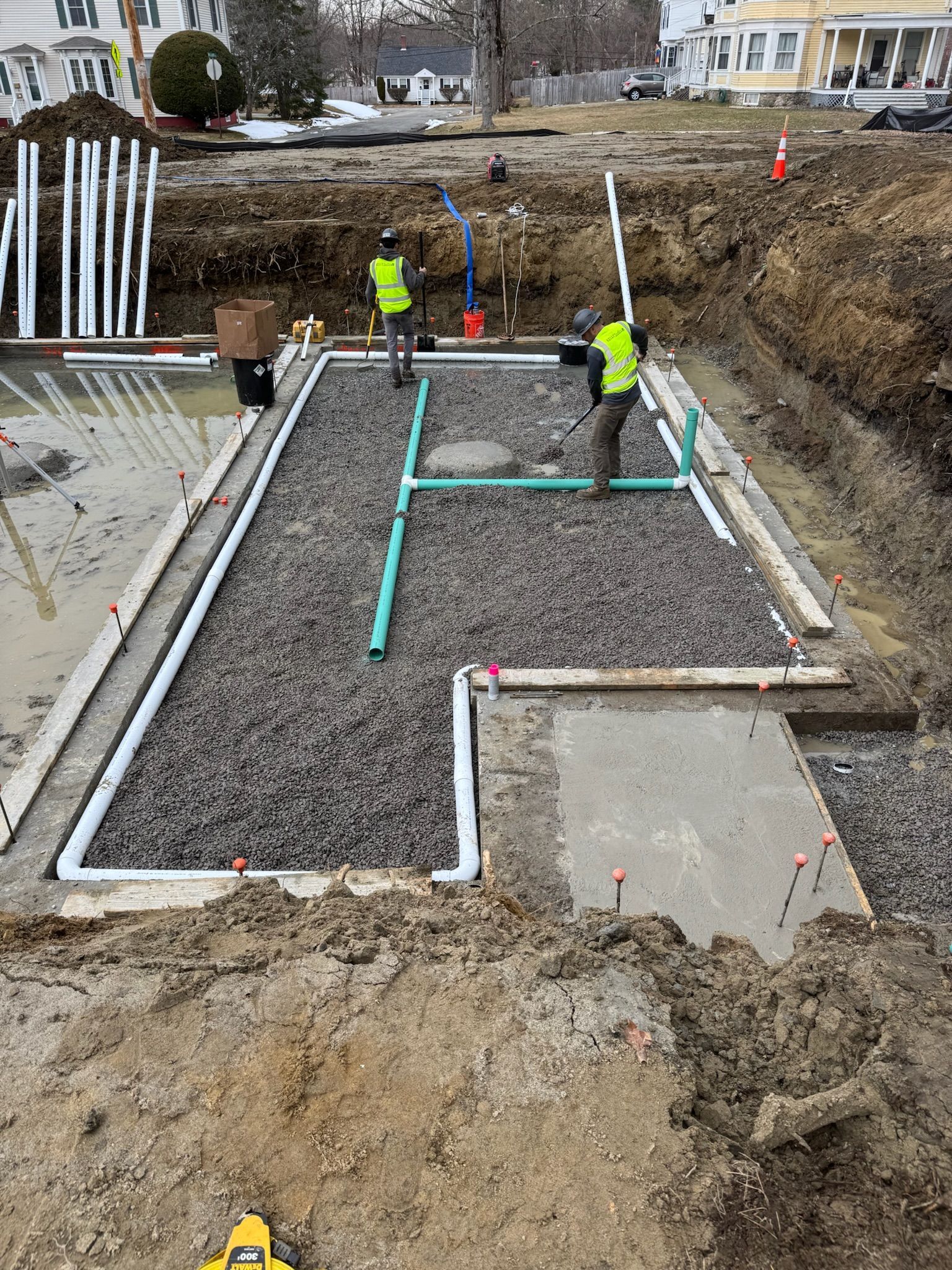 Construction workers inspecting gravel foundation with PVC pipes in an excavated site.