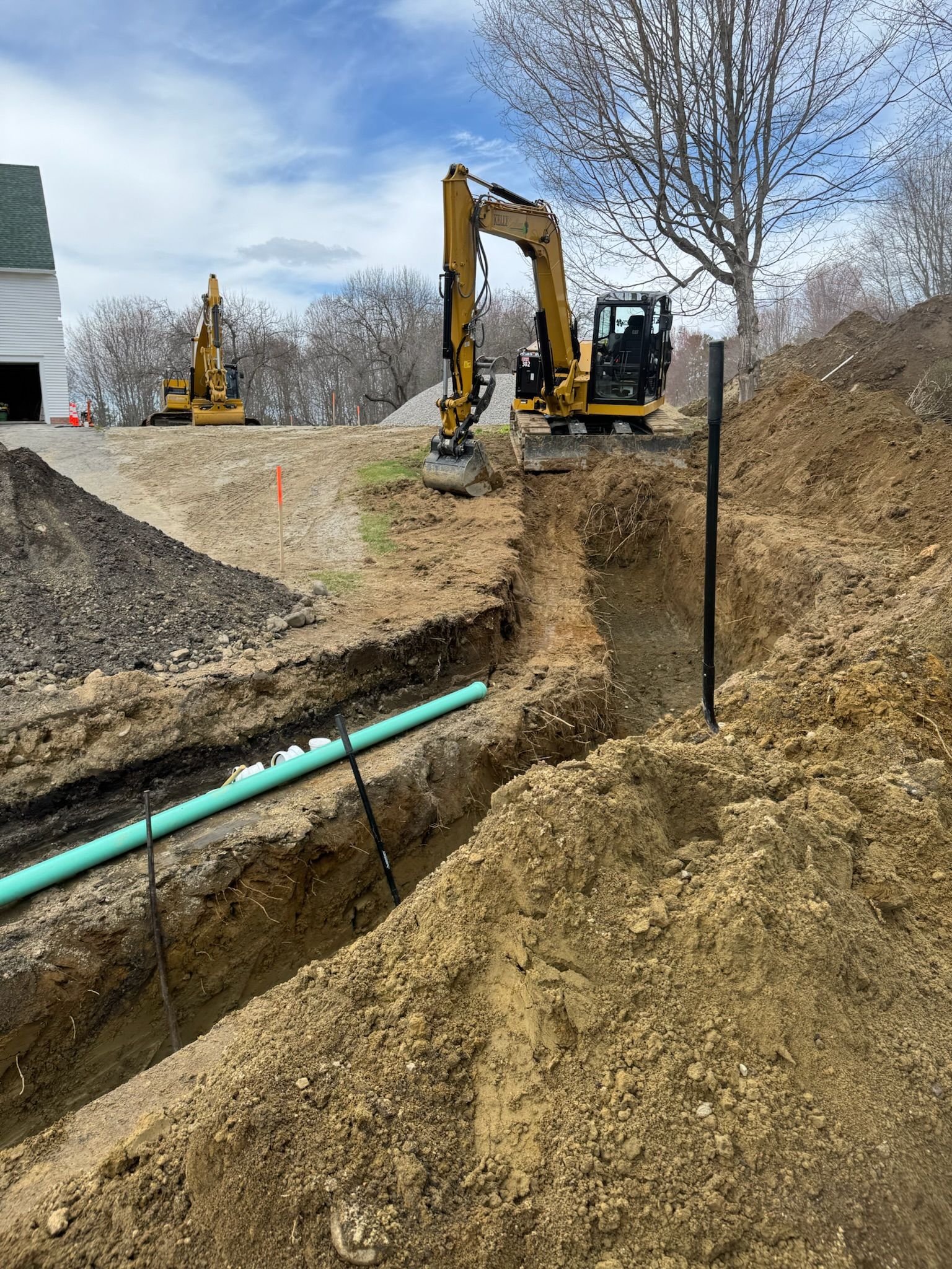 Excavator trenching beside a green pipe at a construction site with dirt piles and bare trees