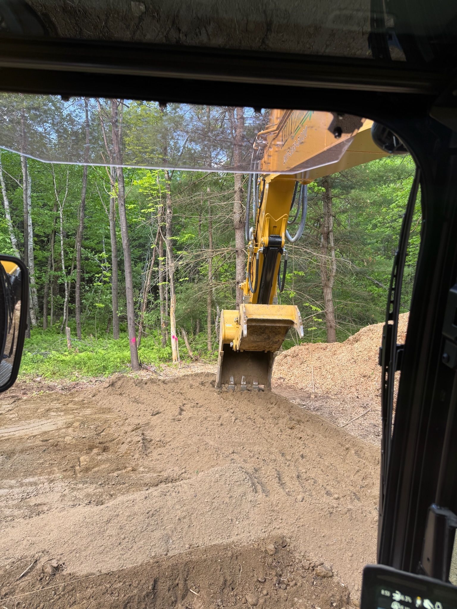 Yellow excavator spreading gravel in a wooded construction area, seen from inside another vehicle.