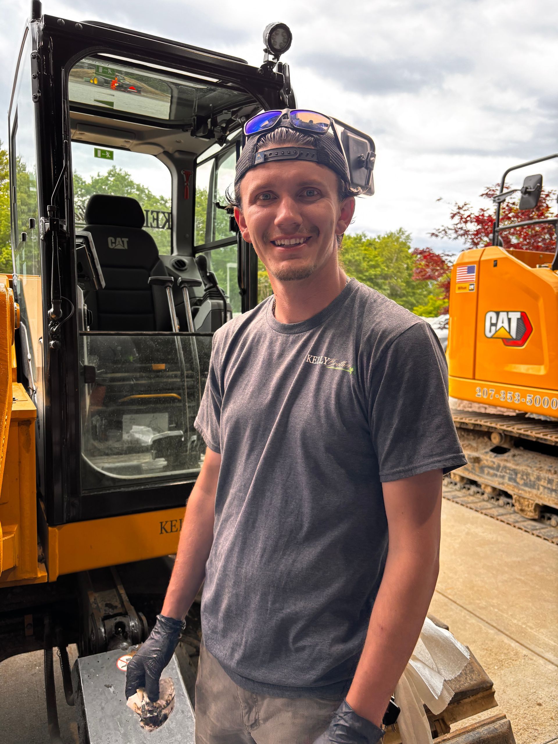 Smiling man in gray shirt and cap standing beside a yellow CAT excavator at a construction site