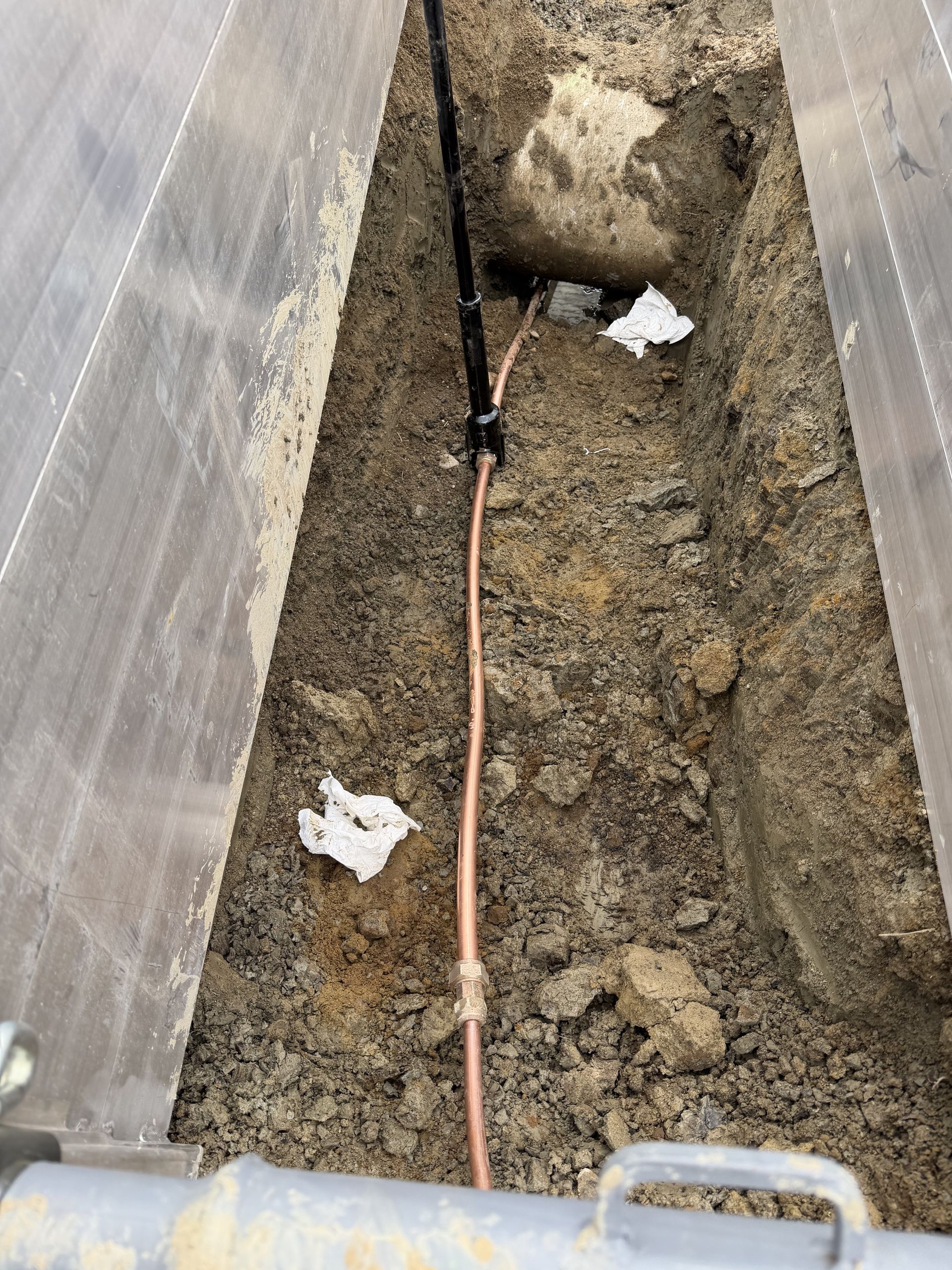 Narrow trench beside a wall with a copper pipe running through dirt and debris