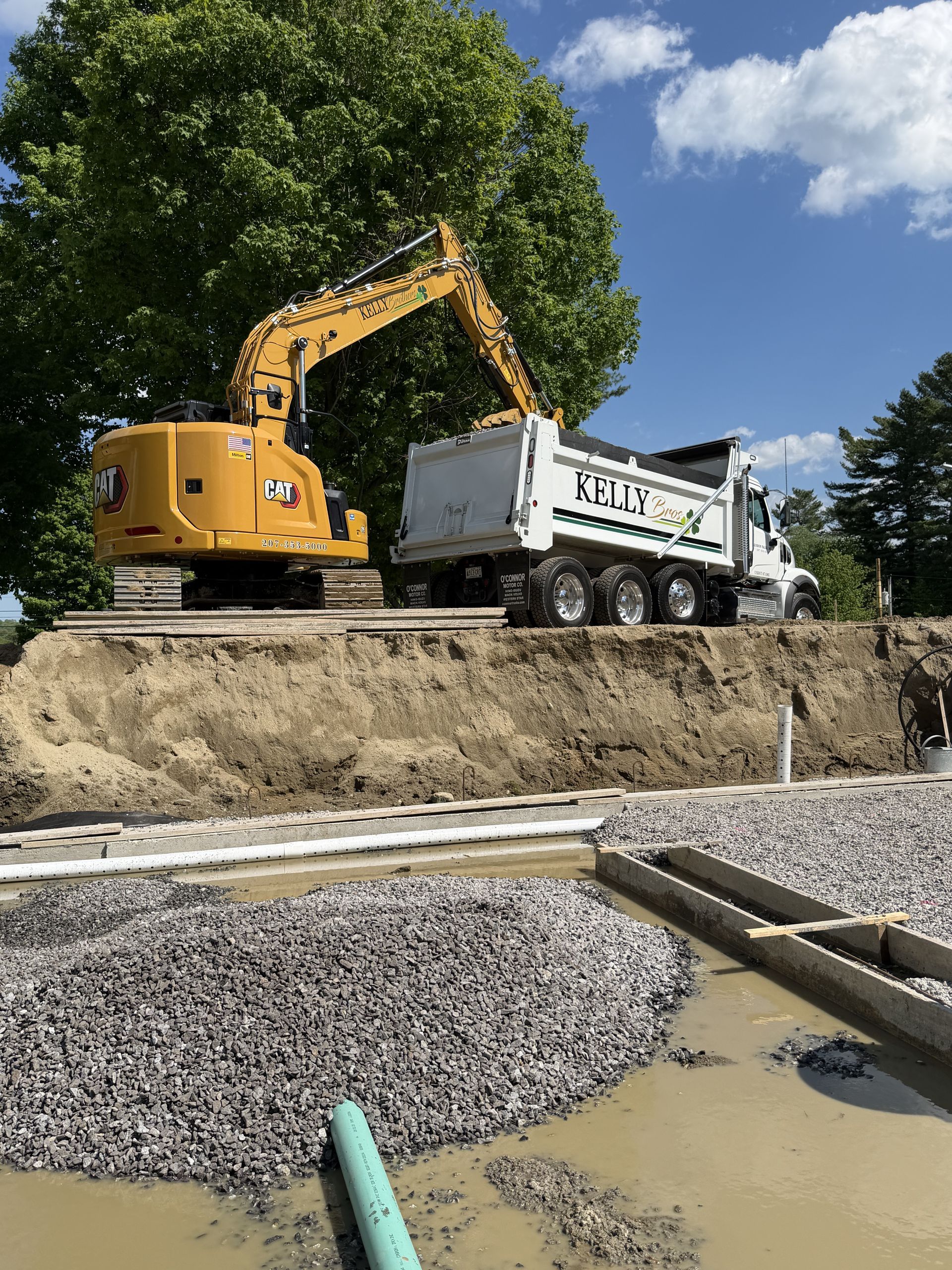 Excavator loading dirt into a white dump truck at a muddy construction site under a blue sky.