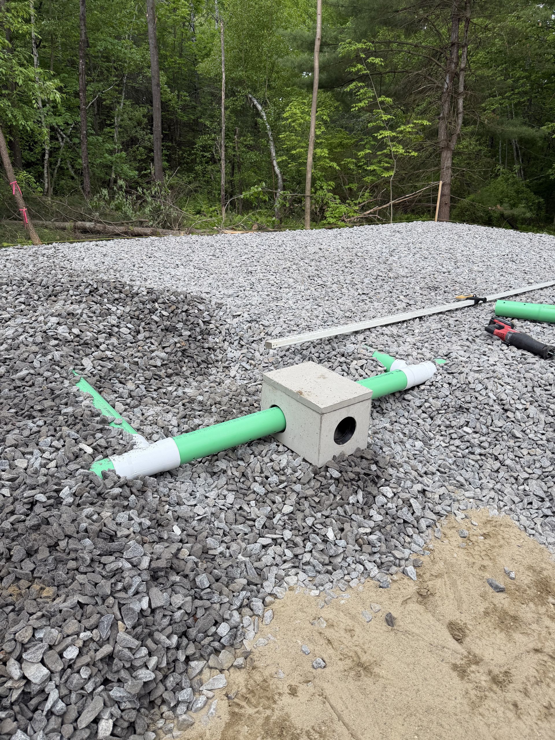 Gravel drainage bed with green PVC pipes and a concrete block, set beside a wooded area
