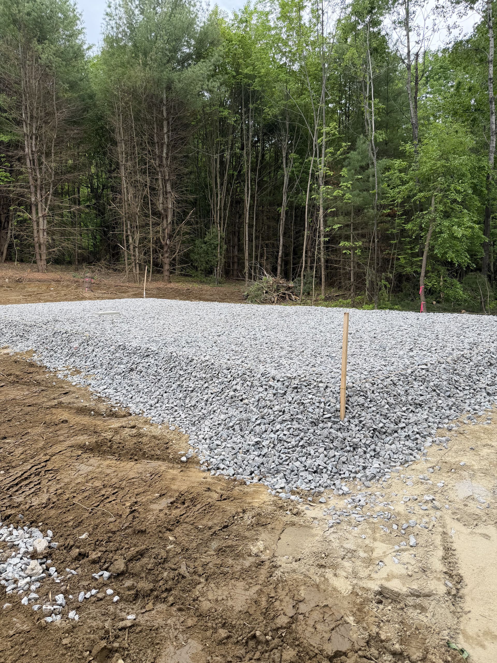 Gravel pad under construction beside a wooded area, with dirt and survey stakes visible.
