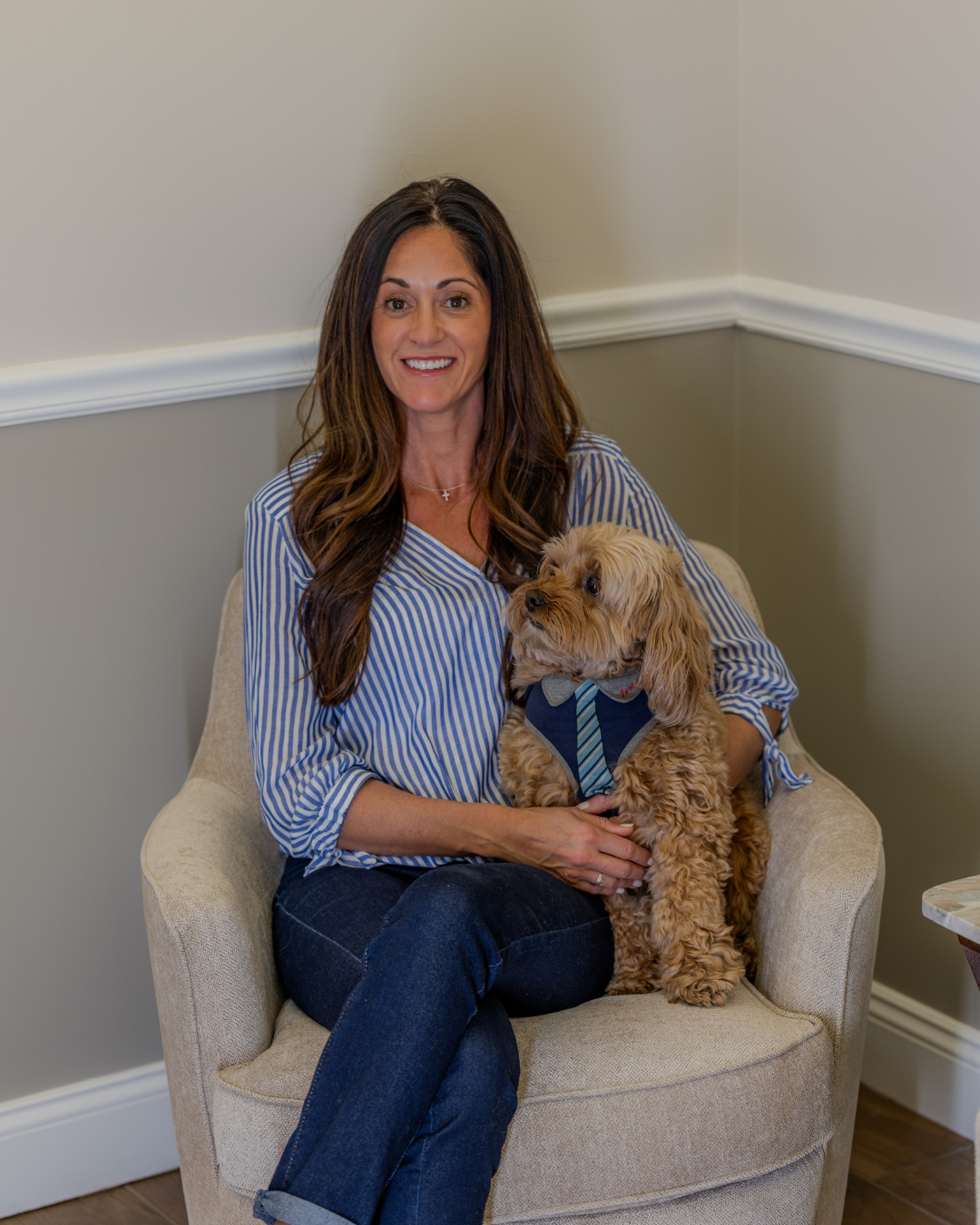 Heidi Kelly seated in an armchair with a small dog in a corner room, both facing the camera.