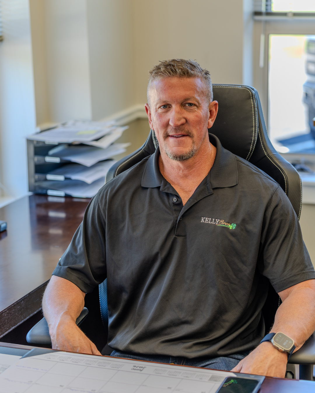 Scott Kelly seated at a desk in an office, wearing a dark polo shirt and smiling at the camera.