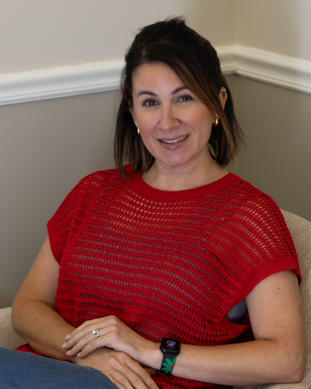 Smiling woman in a red top seated indoors against a beige wall.