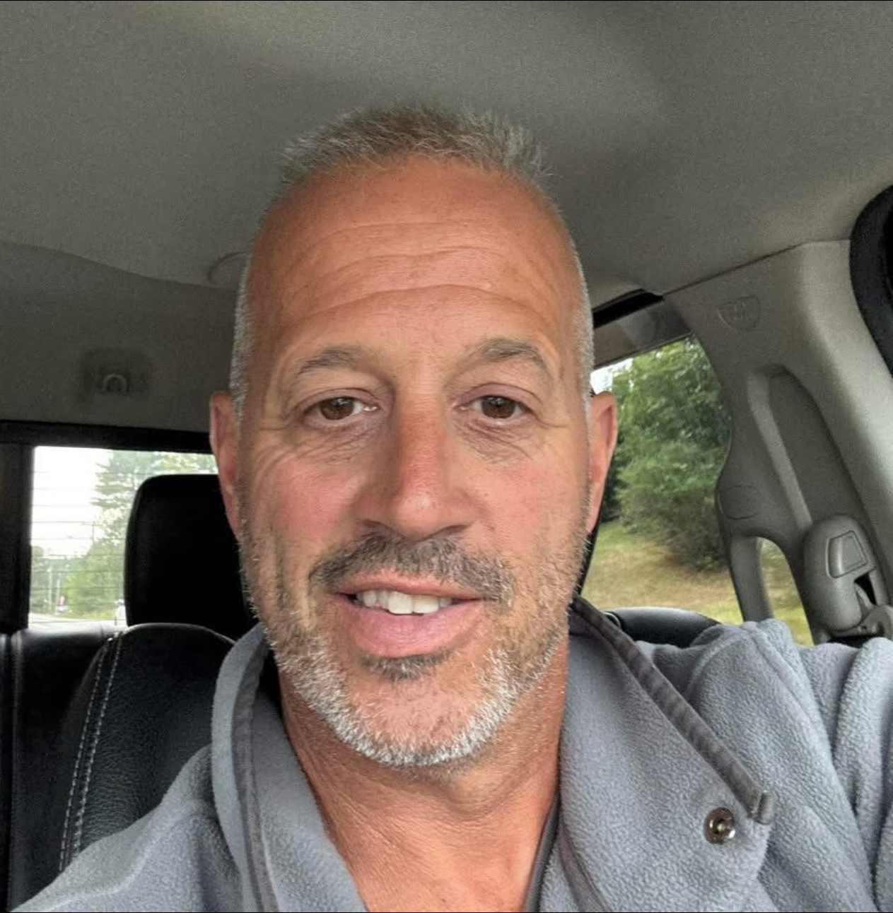 Man smiling in a car, wearing a gray jacket and seatbelt