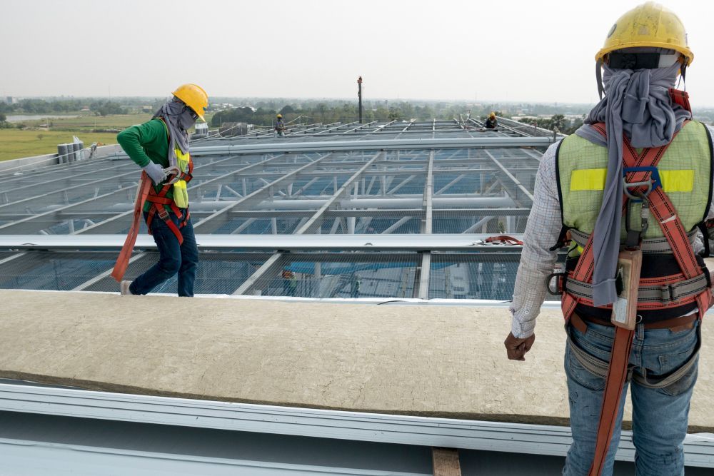 Two construction workers on a rooftop installing metal framing, one holding a green tool.