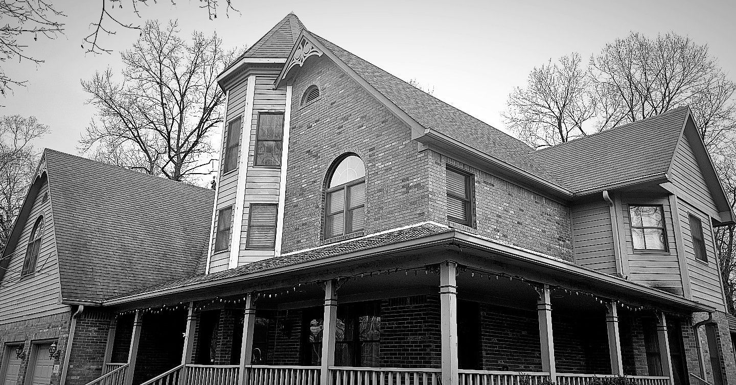 Brick house with steep roof and wraparound porch, viewed from below among bare trees.